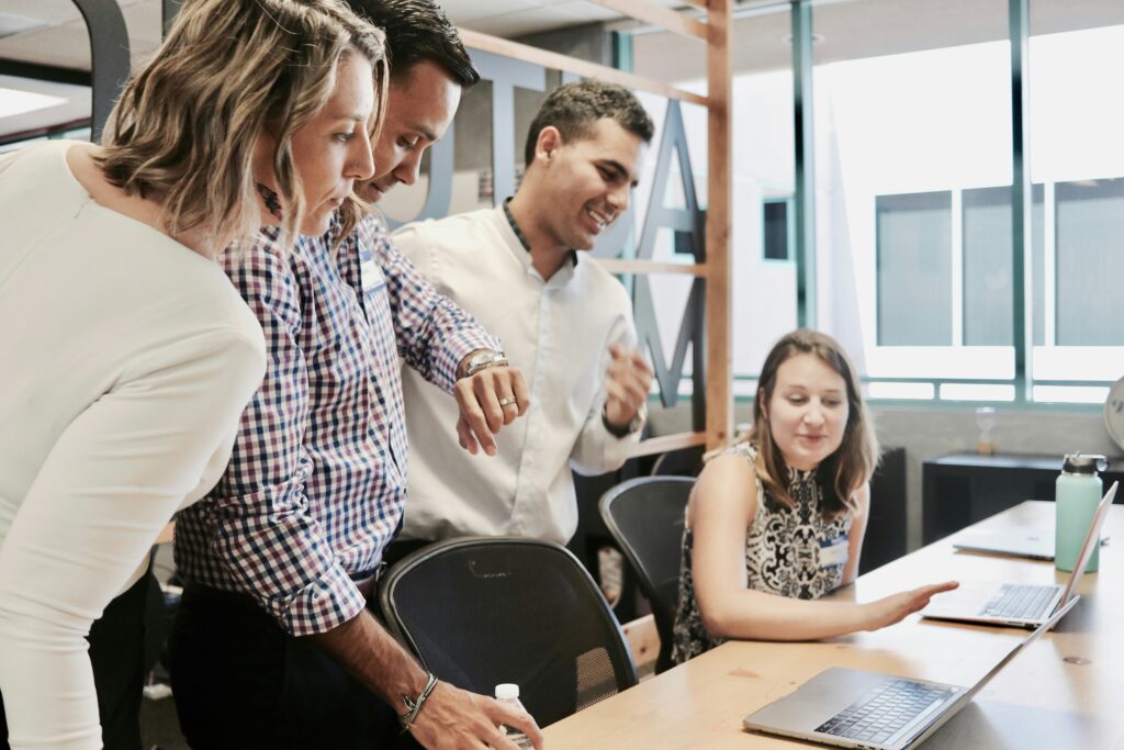 a woman seated at a desk while three people look over her shoulder at a laptop screen
